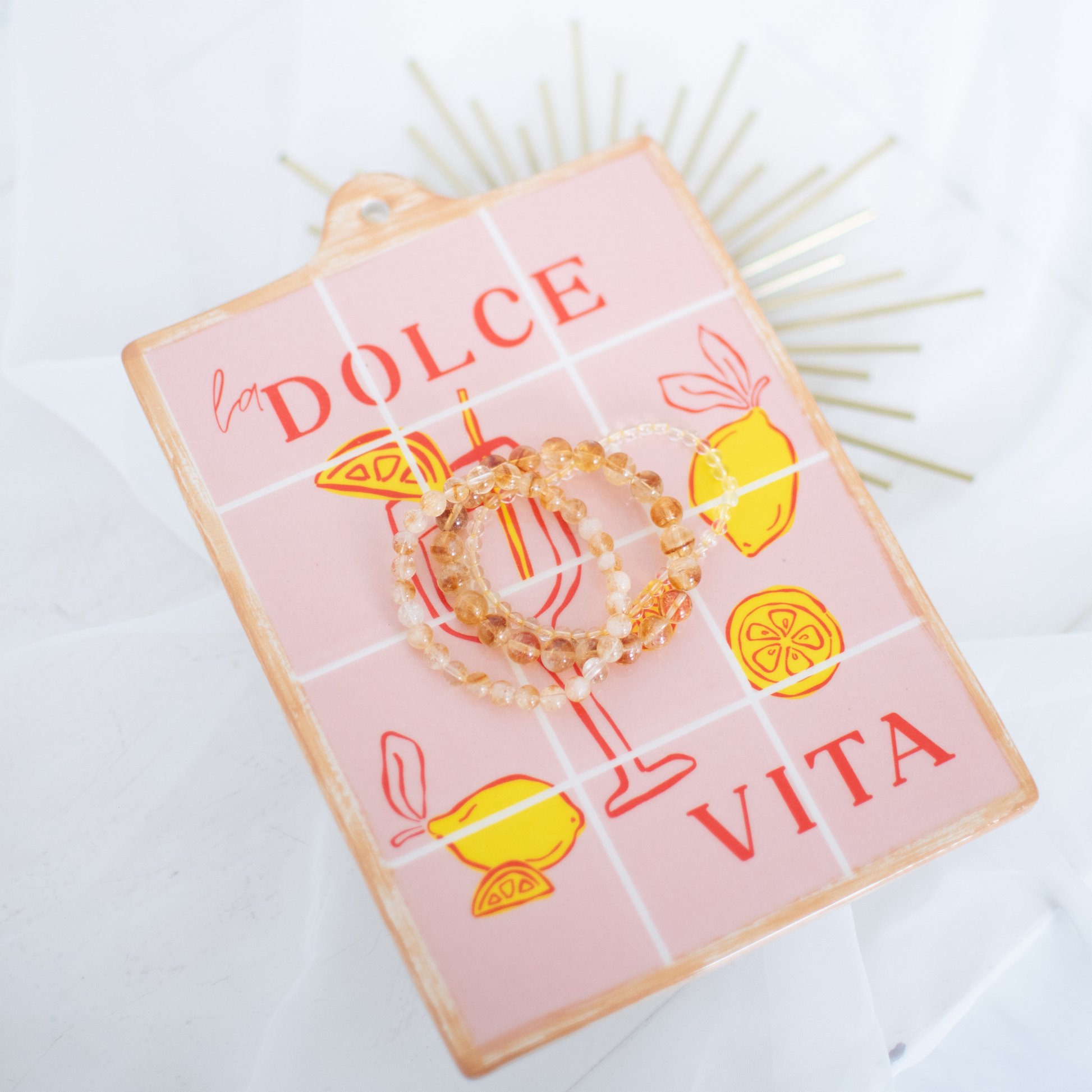 Pink ceramic tile trivet with 'Dolce Vita' text and lemon illustrations on a white background, three beaded bracelets on it