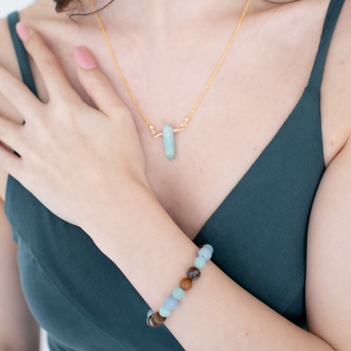 Woman wearing a gold necklace with a turquoise pendant and a matching bracelet on a teal top.
